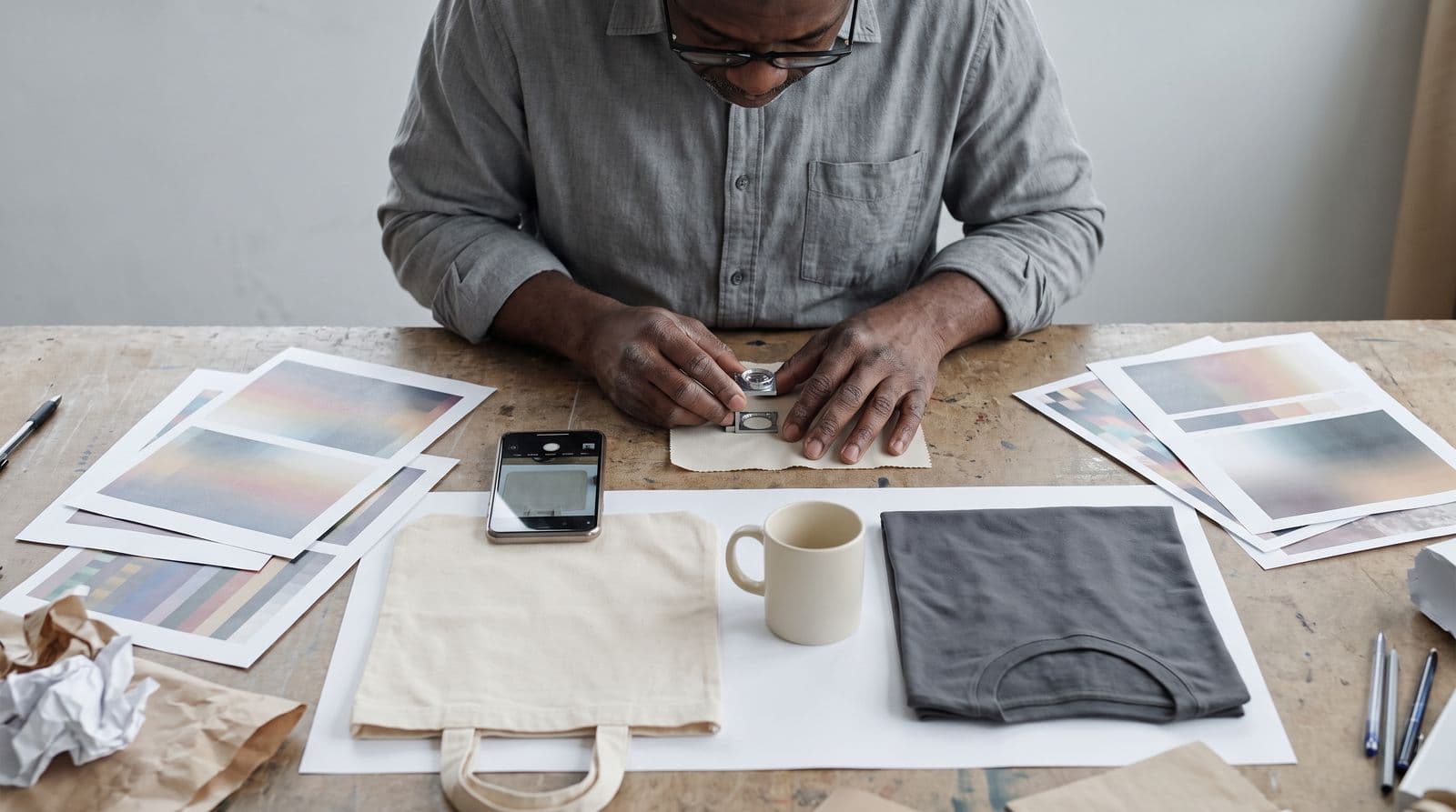 A man in his 50s examines fabric and print quality samples on a neutral work surface using a loupe and a smartphone to compare different production materials.