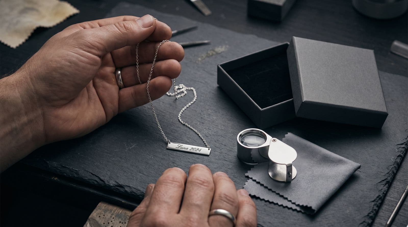 A close-up photograph of a pair of hands holding a sterling silver bar pendant necklace engraved with the text 'Luna 2024' over a dark slate workbench with a jeweler's loupe and an unbranded gift box nearby.