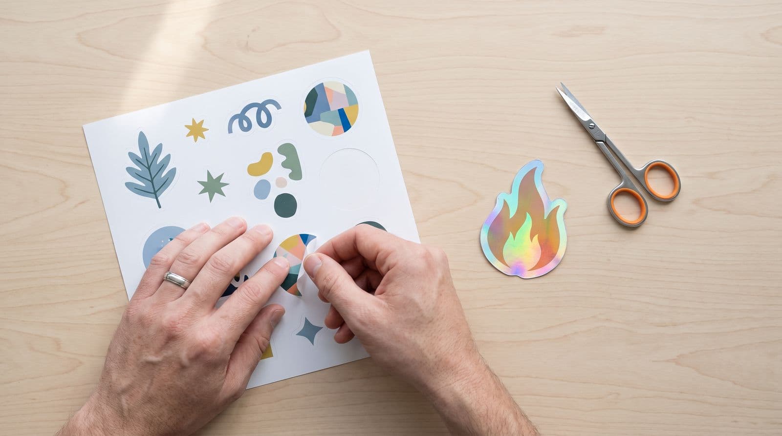 A pair of hands peels a kiss-cut sticker from a sticker sheet on a pale wooden table, next to a holographic die-cut sticker and a pair of scissors.