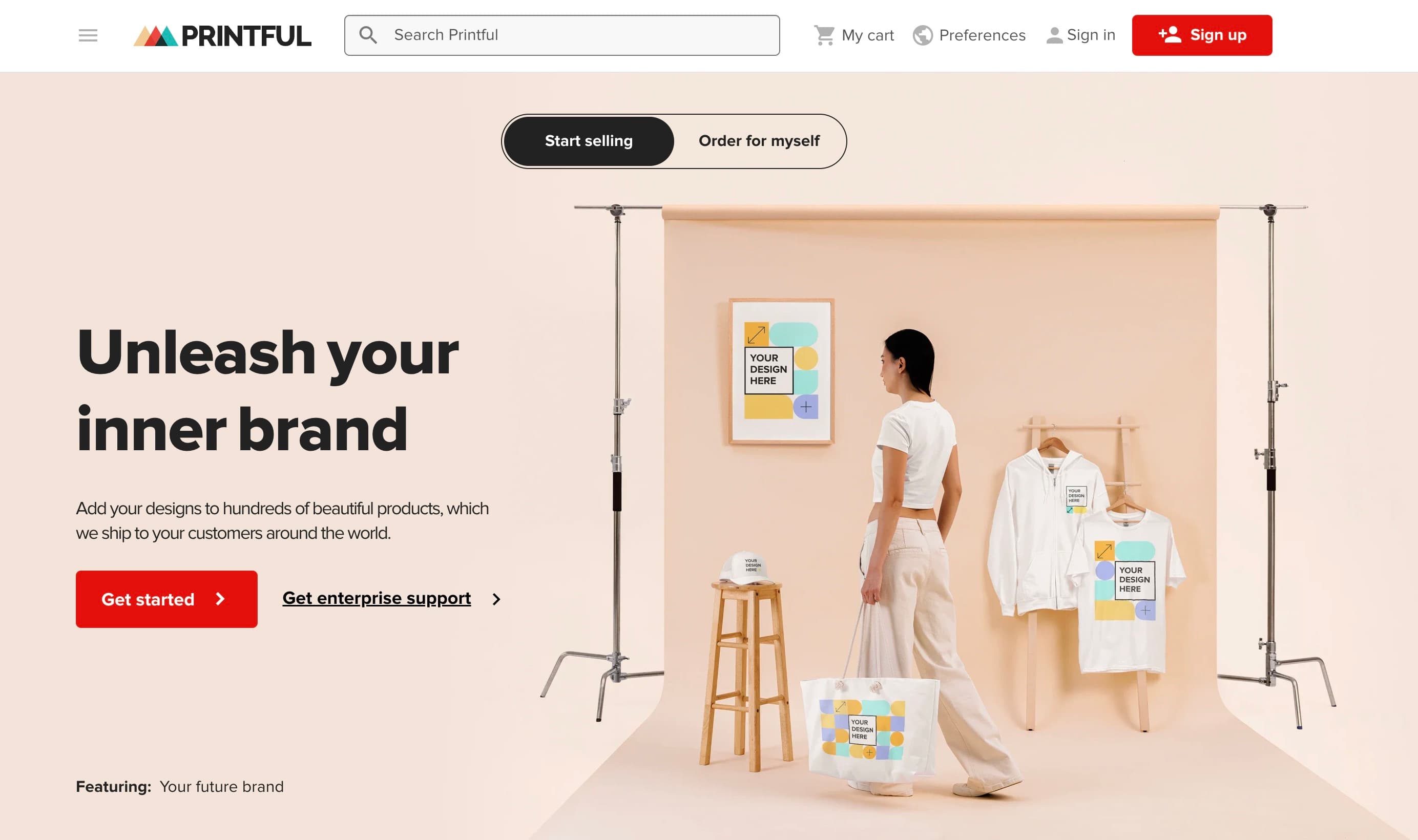 Screenshot of the Printful homepage with the headline 'Unleash your inner brand,' showing a photo studio scene with mockup products including a framed poster, hoodie, t-shirt, and tote bag all labeled 'Your design here.'