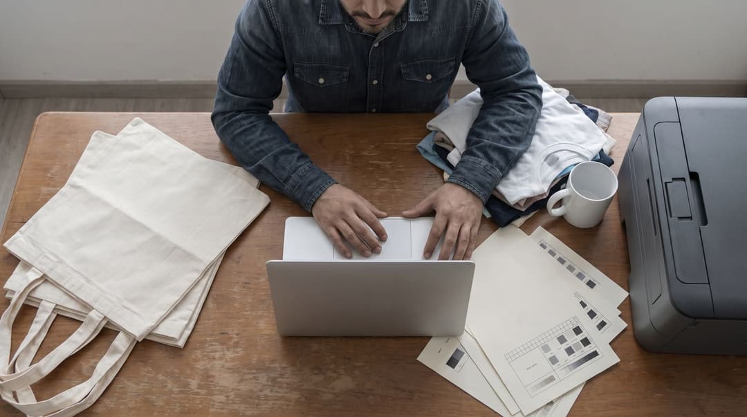 A man in a denim shirt examines print-quality test sheets and various blank apparel samples on a dining table serving as a workspace.