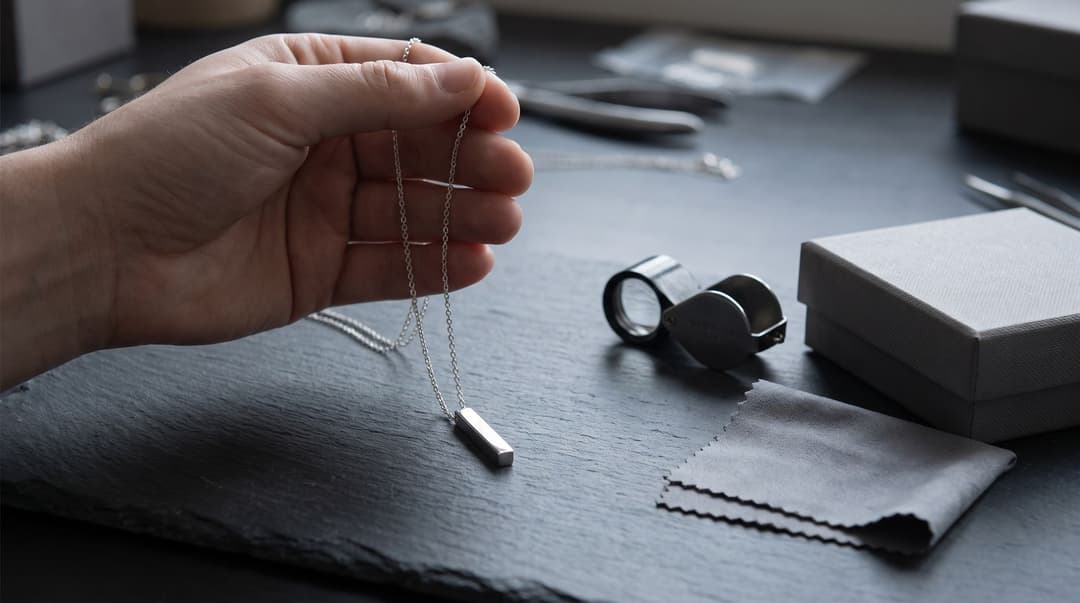 A hand holds a simple bar pendant necklace in front of a dark slate studio bench that features a jeweler's loupe, a cleaning cloth, and a plain gift box.