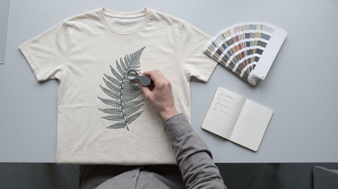 A hand holds a loupe over the fern print on a cream t-shirt, with a colour-swatch fan and a notebook of four ticked checklist lines beside it.