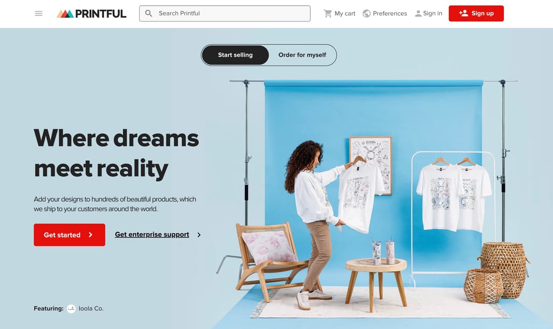 Screenshot of the Printful homepage with the tagline 'Where dreams meet reality,' showing a lifestyle photo of a woman hanging custom-printed t-shirts on a rack.